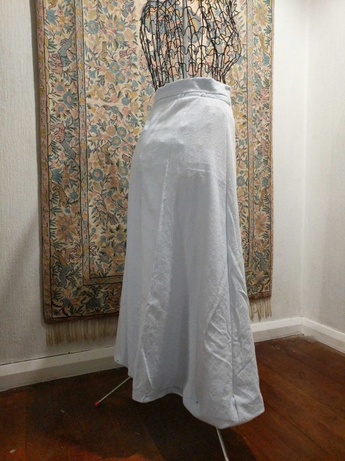 A pale blue flannel petticoat on a wire dressform, in front of an embroidered wall-hanging.