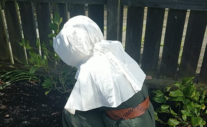 A woman works in a garden with bent head. The sunlight reflects brightly off her white sunbonnet.