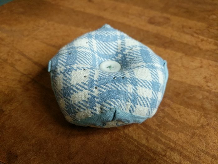 A tubby little biscornu pincushion of checked flannel, dimpled in the middle with a white button. There are a scattering of silver pins. The blue bias binding zig-zagging around the circumference mediates between the checked fabric and the yellower floral that can be glimpsed at its base.