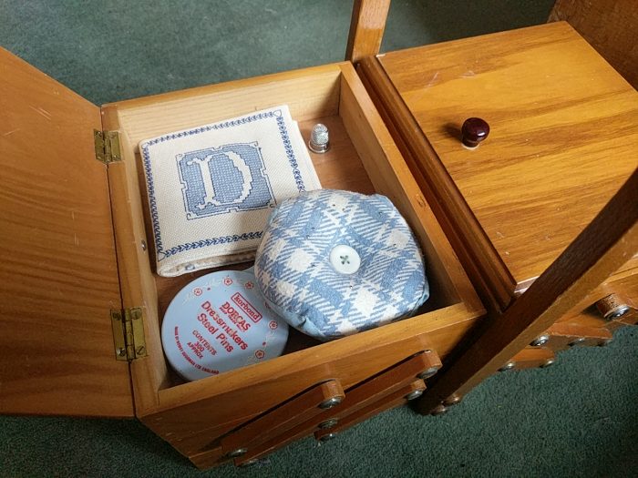 A concertina sewing box with one side open. Inside are a blue-and-cream biscornu pincushion, a blue-and-cream embroidered needlebook, a blue-lidded container of pins, and a silver thimble.