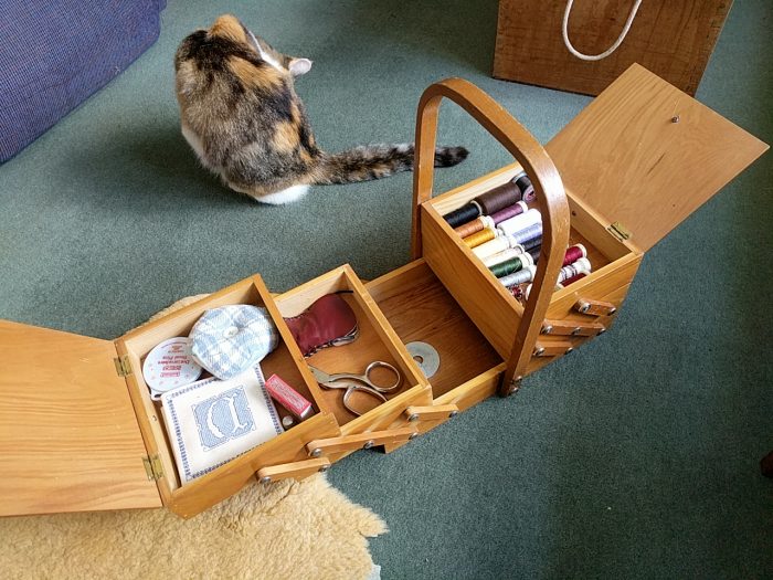 A sewing box shown with both lids open and one side extended. Inside are a range of threads, scissors, and other sewing equipment. In the background a cat licking itself.