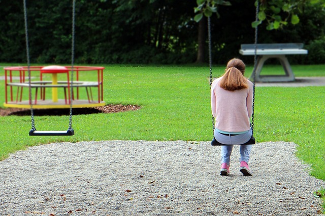 A woman sits sadly on a swing, back to the viewer. The swing next to her is empty. In the background is a roundabout, also empty.