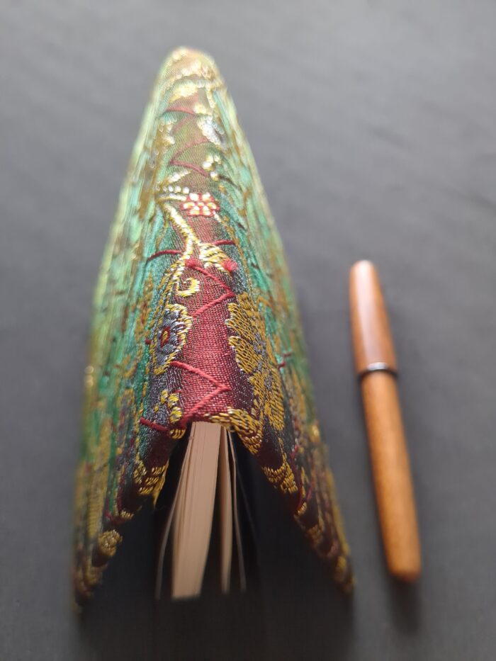 A small fabric-covered book sitting slightly open on its open edge. The near part of the spine appears red, but the further part and the covers appear to be a mix of green tones.