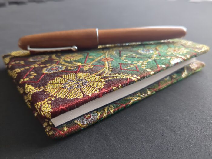 A brown wooden fountain pen resting on a small notebook with a red/green shot silk cover ornamented with gold.