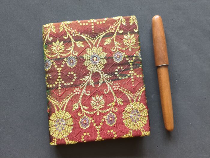 A small book covered in a red fabric with quantities of stylized floral ornamentation in gold and silver. Next to it, a fountain pen in brown wood.