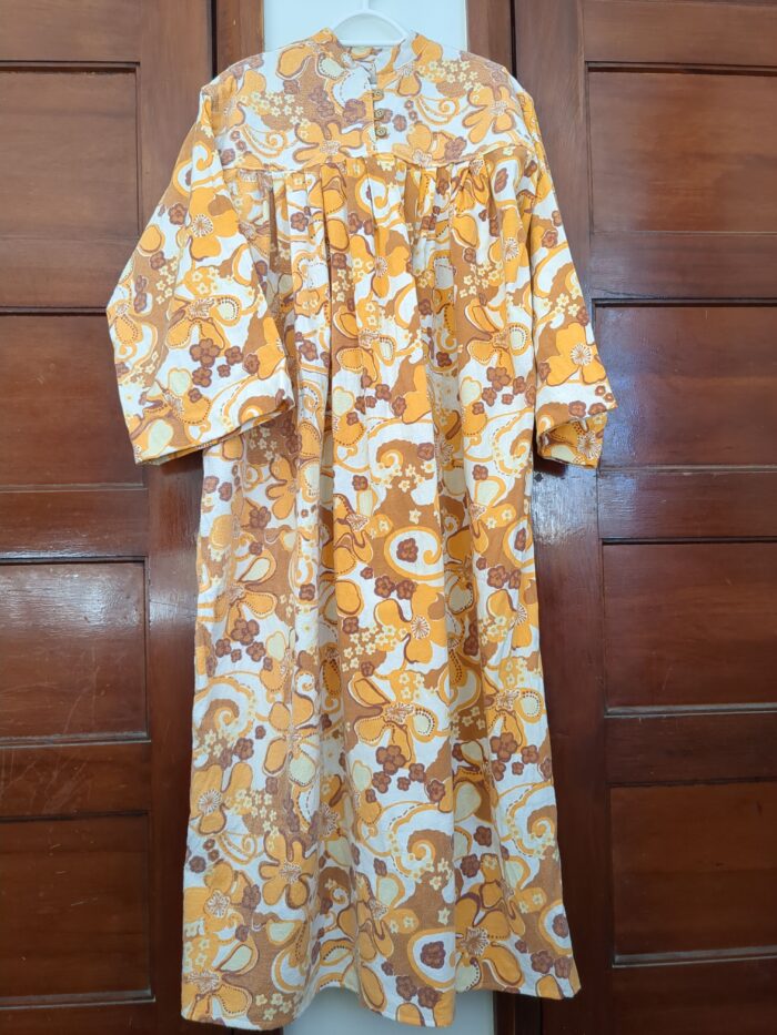 A full length winter nightgown hanging on a coathanger between two wooden doors. The nightgown has long ungathered sleeves, and pleats hanging from a buttoned yoke. The pleats are hard to distinguish due to the loud retro pattern of the fabric.