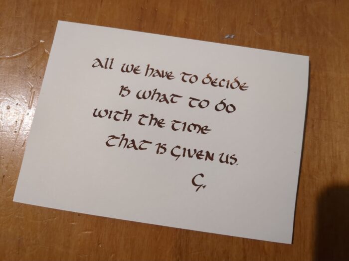 A sheet of paper on a wooden surface has uncial lettering in brown ink which reads "All we have to decide is what to do with the time that is given us. G."