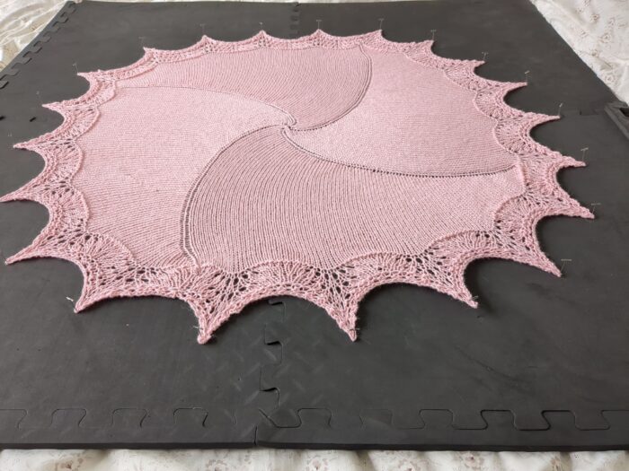 A circularish pink shawl pinned out to dry on dark grey foam boards. Four lines of increases spiral out from the middle, and the edging is of spiky lace.
