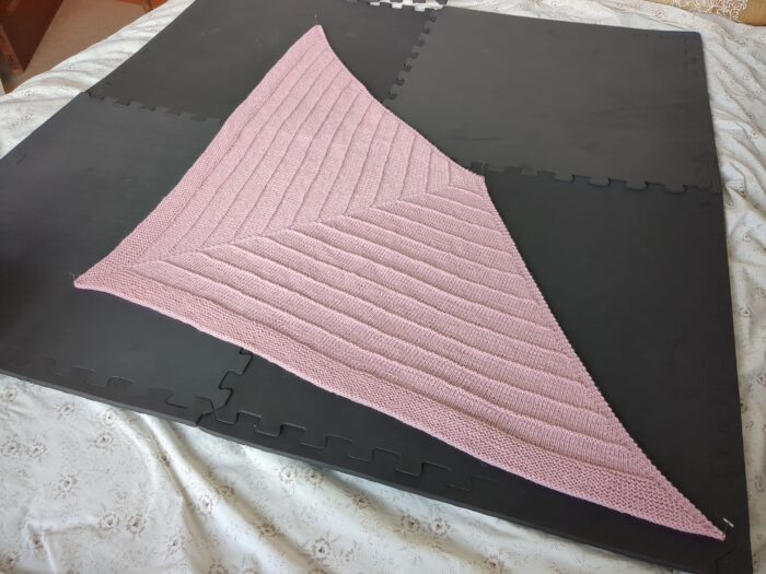 A more or less triangular pink shawl spread out to dry on dark grey foam boards. The shawl is largely smooth stocking stitch, with bands of garter stitch standing out to give it texture.