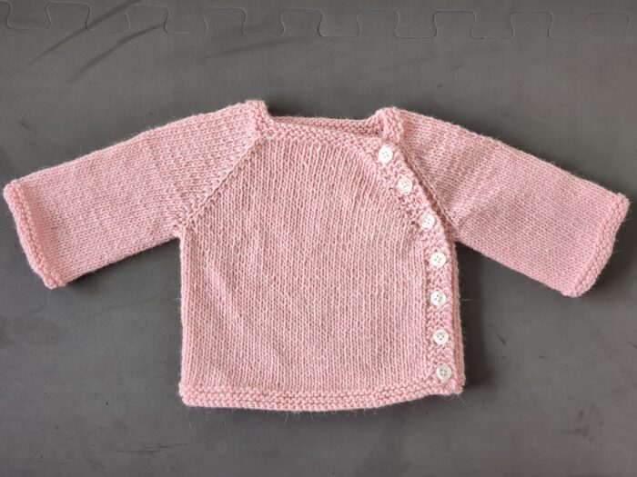 A small pink 8ply baby cardigan with seven buttons running from neckline to underarm to hem down the side of the garment.
