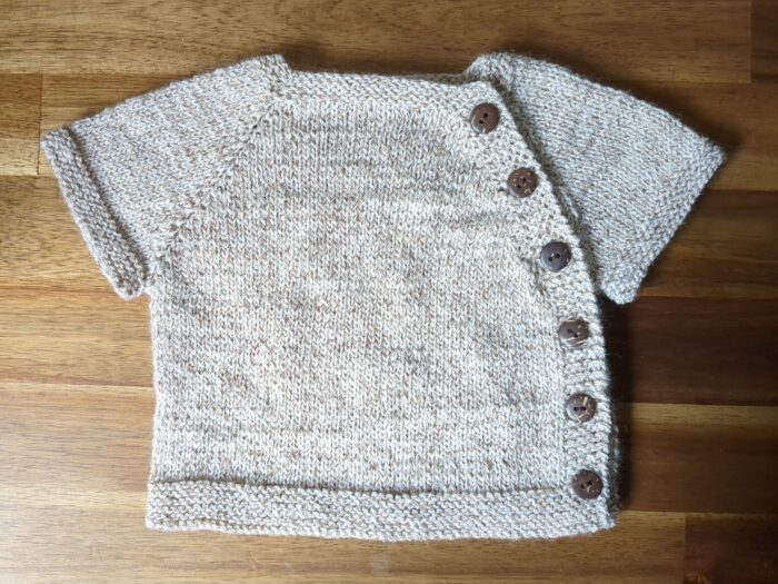 A porridge-coloured short sleeved baby cardigan with six coconut shell buttons running from neckline to underarm to hem down the side of the garment.
