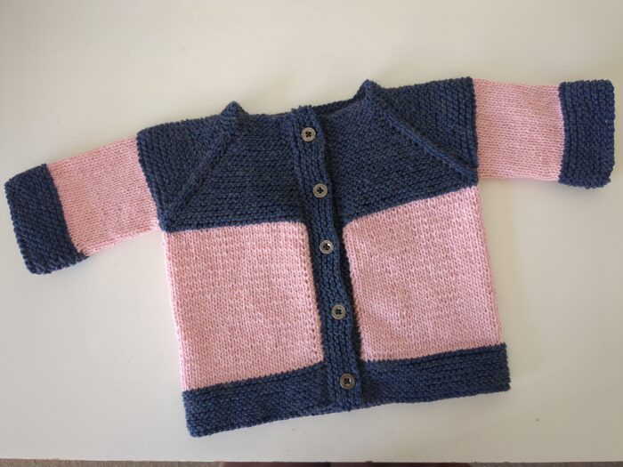 A small 8ply baby cardigan in pink with dark blue garter stitch trim and buttons down the front.