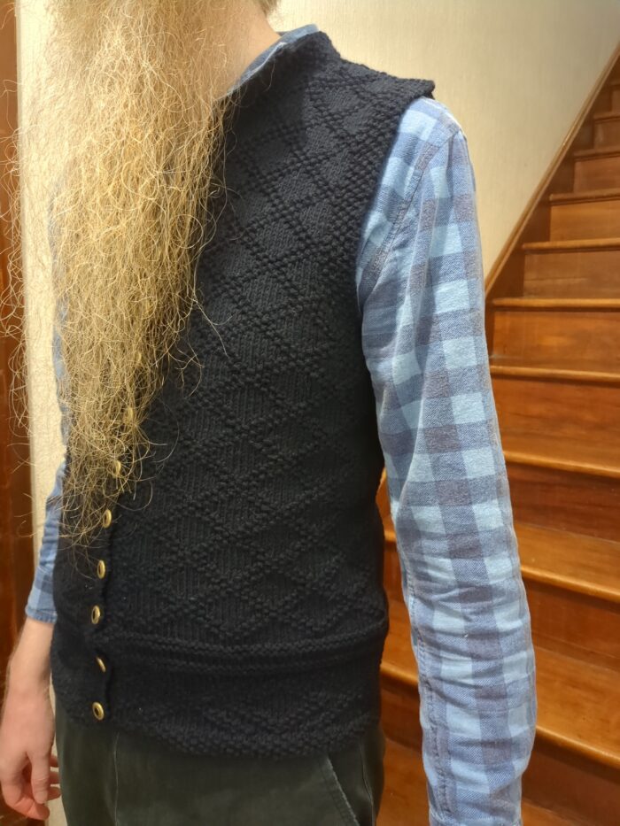 The left side of a dark blue knitted waistcoat with a diamond lattice pattern made of purl stitches, worn by a man with a long orange beard.