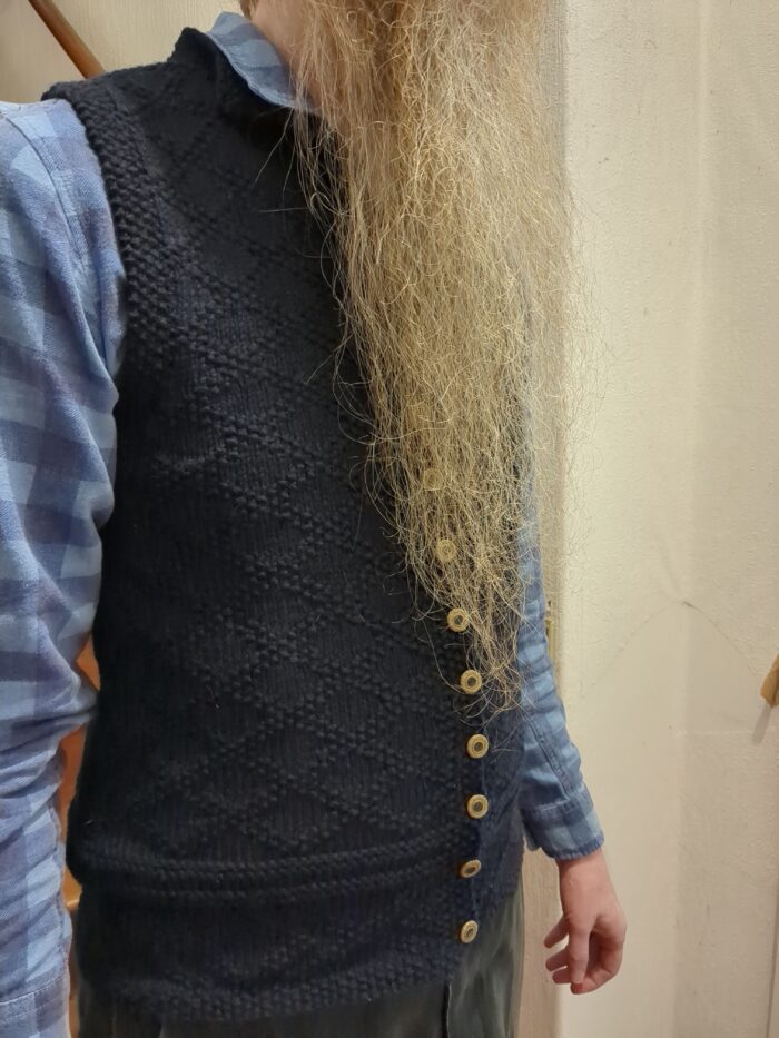 The right side of a dark blue knitted waistcoat with a diamond lattice pattern made of purl stitches, worn by a man with a long orange beard.