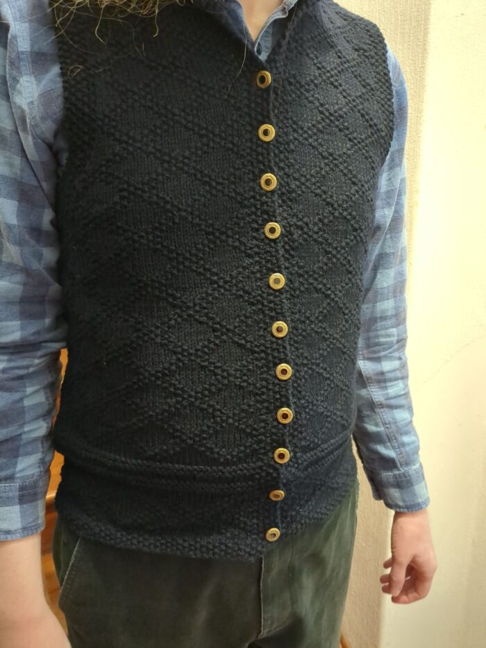 A dark blue knitted waistcoat with a diamond lattice pattern made of purl stitches. It has eleven buttons down the front.