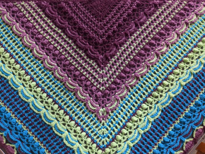 The central part of a large triangular shawl. A repeating pattern of netting, bobbles, and swags changes colour with each repeat. The first repeat is dark purple and berry pink, the second berry pink and light green, the third light green and bright blue, and the fourth bright blue and dark purple.
