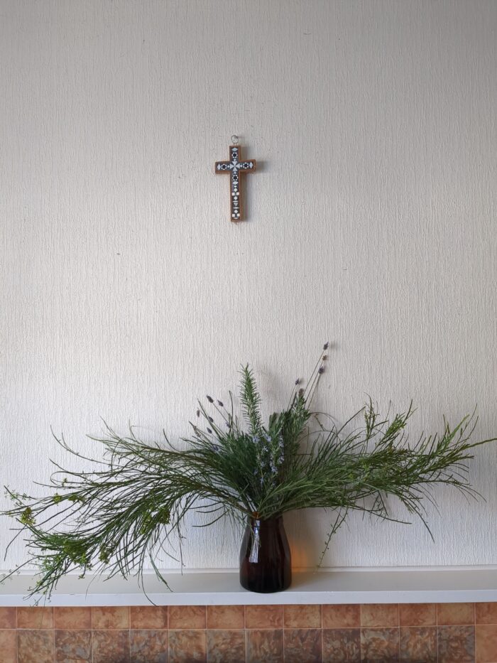 A decorative mosaic cross hangs on a textured off-white wall above a white mantleshelf over terracotta tiling. On the shelf is a large brown glass jar with a flower arrangement. Long branchy arms swoop out to left and right, with spires of lavender and rosemary rising in the centre.
