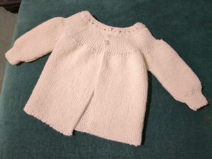 A cream wool baby jacket, with two little buttons fastening a yoke below an eyeleted collar.