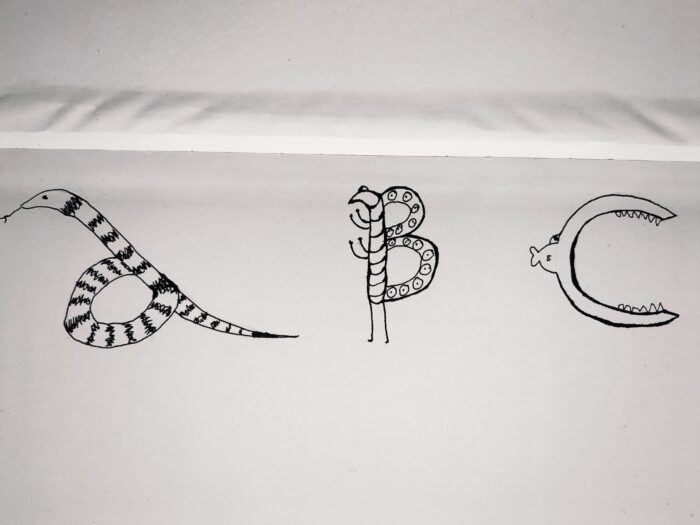 Three amateurish drawings in black ink in a sketchbook. From left to right: a stripy snake coiled into the rough shape of a lower case a; a rather cartoonish butterfly with striped body and patterned wings forming the shape of an upper case letter B, standing on two spindly legs with two other pairs raised in a pugilistic manner; a tiny fish with an enormous open mouth in the shape of the letter c.
